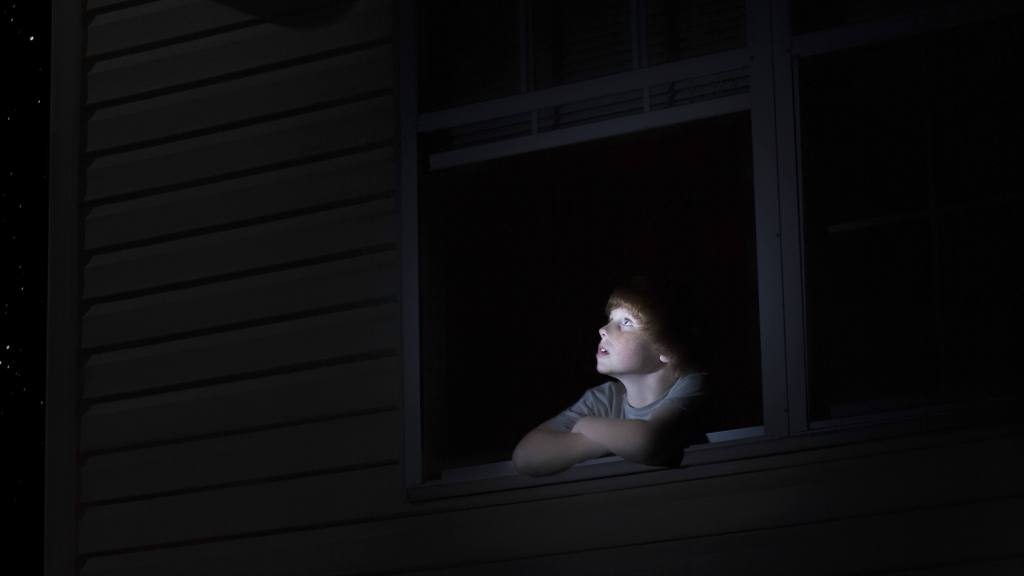 A boy gazes at the stars through a darkened window at night.
